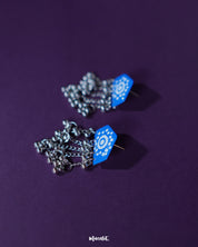 Nazafar blue jhumkas presented front-facing on a simple backdrop, showcasing the striking white circular artwork while highlighting the oxidized silver dome and cultural elegance – Muselot.