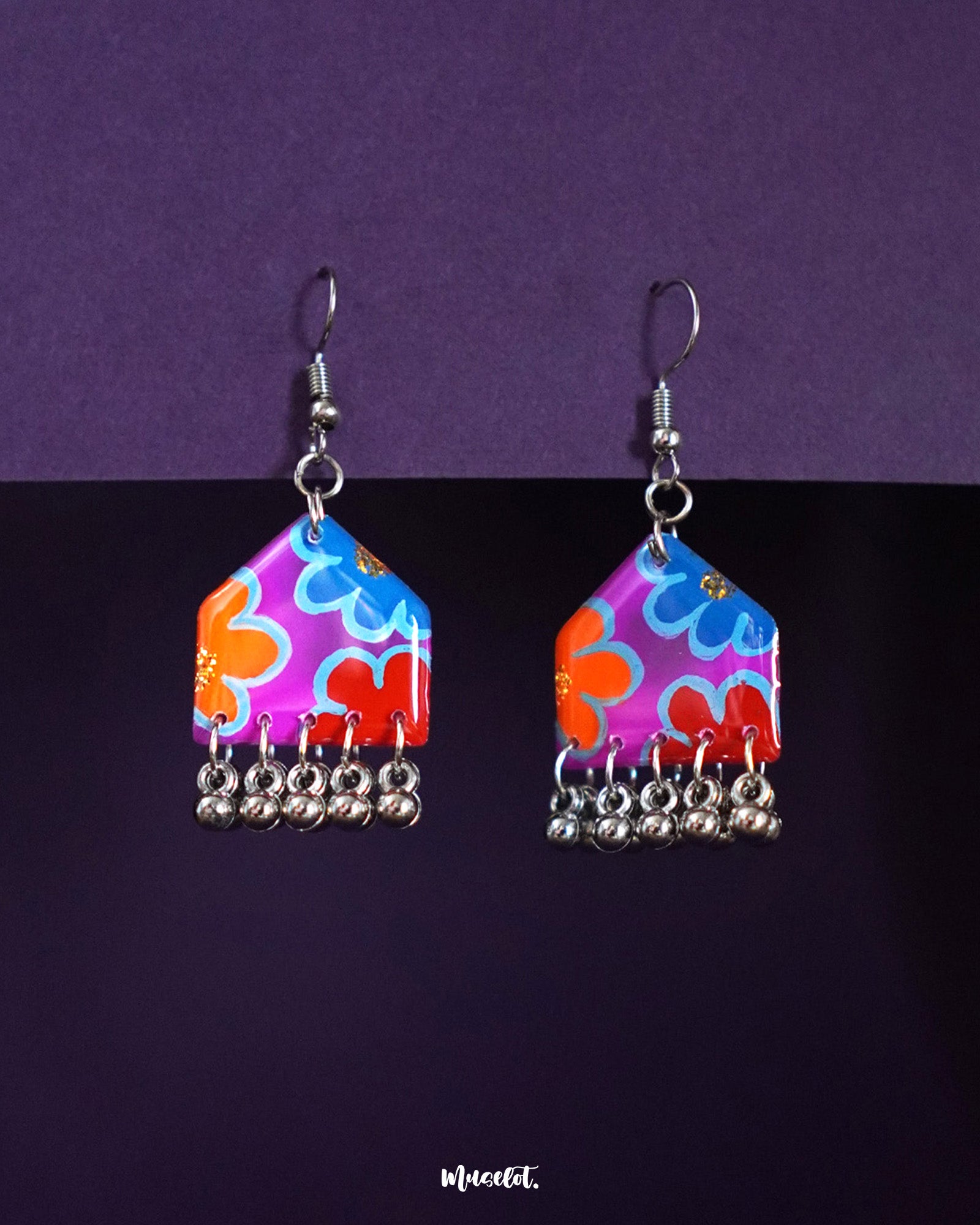 Nazrana hand-painted hook earrings with multicolour floral motifs on a purple-red base and silver ghungroo drops, displayed against a deep purple background – Muselot.