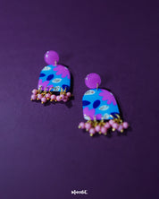 Close-up of Neelzair purple floral earrings featuring hand-painted pink and blue florals with delicate golden specks, highlighting the artisanal detailing and showcasing the charm of the pink beaded danglers — Muselot.