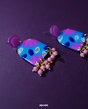 The Neelzair purple floral earrings rest against a purple background, highlighting their vivid color palette and intricate floral design with beaded fringes — Muselot.