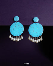 Neerangi pearl earrings featuring small and large round turquoise blue discs embellished with subtle silver glitter accents, finished with dangling white pearl-like beads, beautifully displayed against a deep purple background – Muselot.
