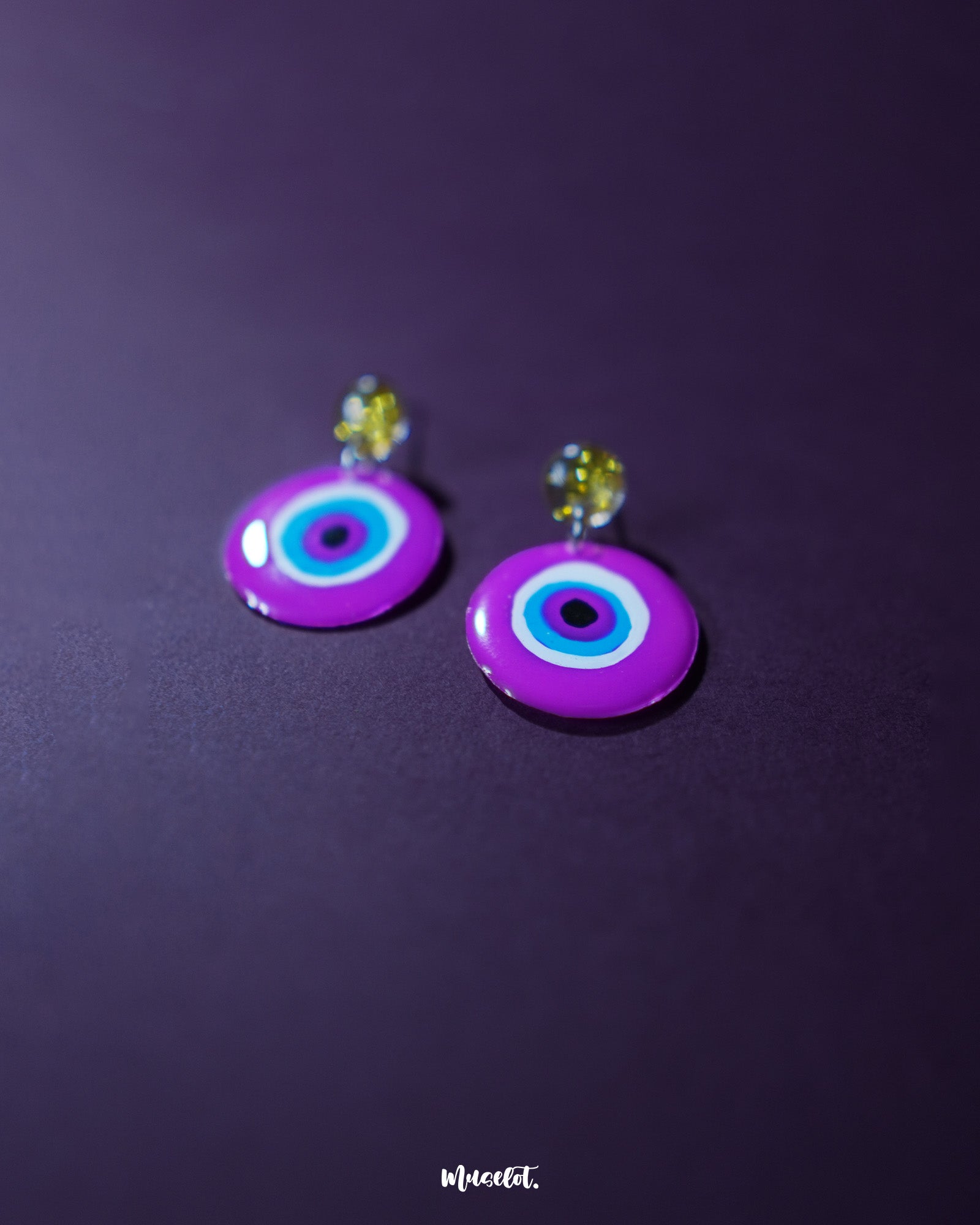 Nigaahbaan evil eye stud earrings rest elegantly against a rich purple backdrop, with their vibrant magenta discs and hypnotic concentric circles drawing in all eyes—radiating protection with a pop of playful color — Muselot.