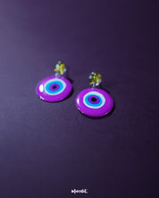 Nigaahbaan evil eye stud earrings rest elegantly against a rich purple backdrop, with their vibrant magenta discs and hypnotic concentric circles drawing in all eyes—radiating protection with a pop of playful color — Muselot.