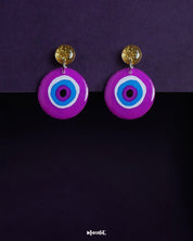 A bold and playful pair of Nigaahbaan stud earrings, featuring a hand-painted evil eye motif in vivid purples and blues, anchored by shimmering golden studs—perfect for warding off the ordinary — Muselot.