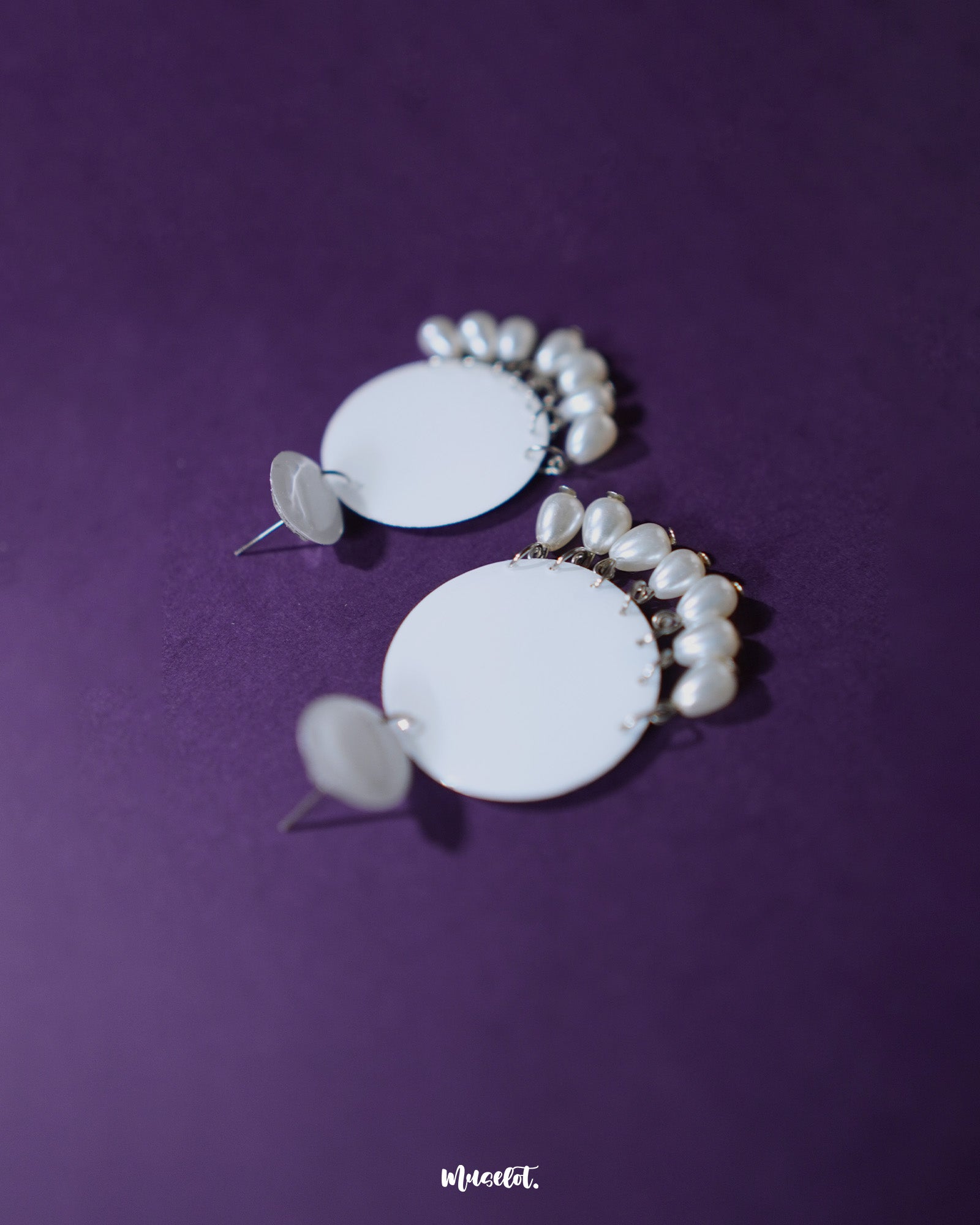 Noor white drop earrings displayed against a deep purple background, featuring glossy round tops with matching circular drops adorned with elegant pearl hangings, showcasing minimal elegance and highlighting timeless charm — Muselot.