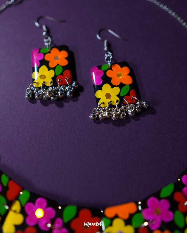 A focused shot of the earrings from the Parizad floral jewelry set, featuring vibrant hand-painted flowers in pink, yellow, red, and orange on a black base. The silver gungroo drops at the bottom add movement and charm, making them perfect for a playful yet artsy boho look — Muselot