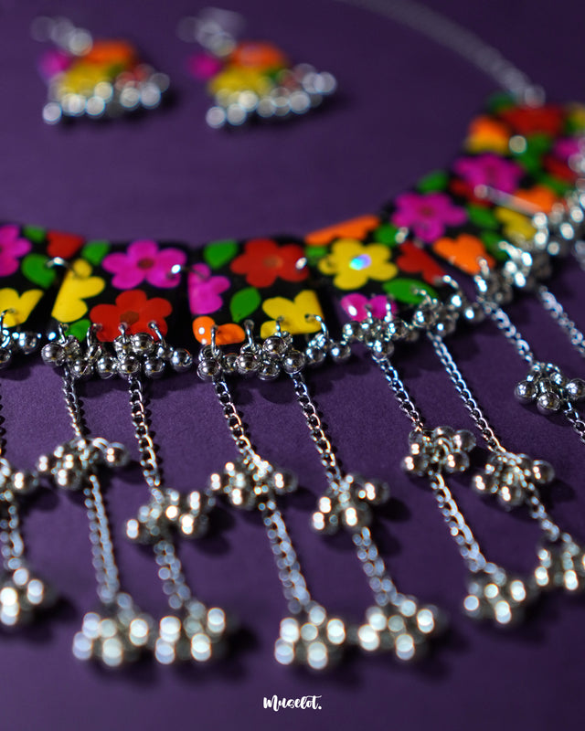 A vibrant close-up of the Parizad floral jewelry set laid out on a deep purple background, showcasing its hand-painted multicolor floral design on a black base. The playful silver gungroo drops and cascading chains highlight the set’s handcrafted boho charm — Muselot.