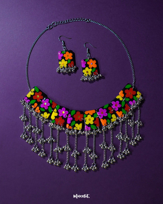 A vibrant floral jewelry set featuring a necklace and matching earrings, laid flat on a rich purple background. The black base is adorned with colorful hand-painted flowers in pink, orange, yellow, green, and red, finished with cascading silver gungroo chains. The piece showcases a bold boho style, perfect for uplifting everyday looks — Muselot