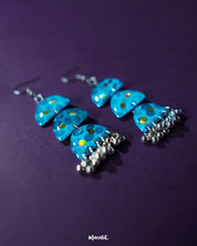 A pair of Parvaaz blue drop earrings displayed on a soft purple background, showcasing their vivid hand-painted blue floral design and elegant drop silhouette that reflects traditional craftsmanship with a modern charm — Muselot.