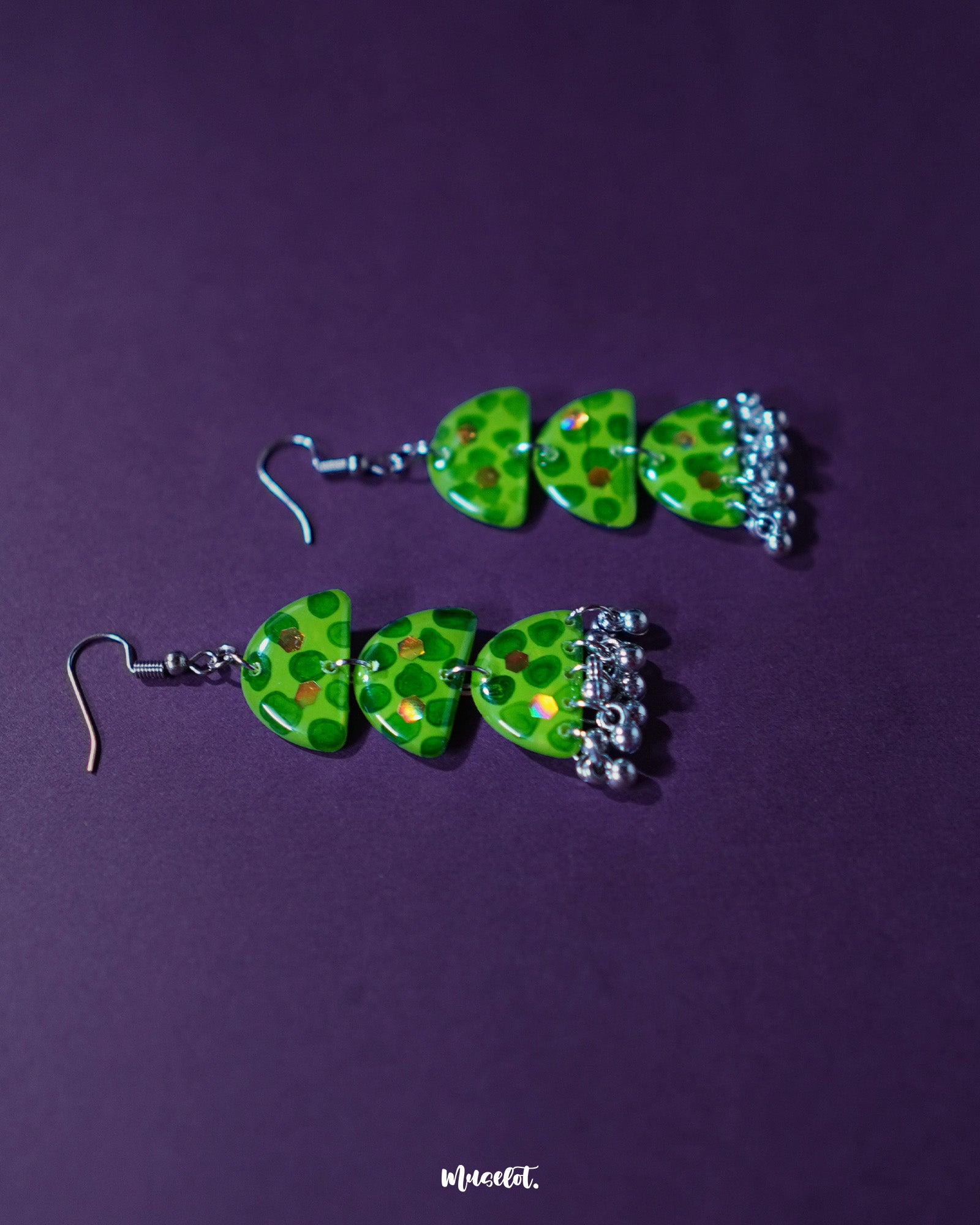 A pair of Parvaaz green drop earrings displayed against a soft pastel background, featuring deep green dotted accents on a light green base. The teardrop design highlights the delicate craftsmanship and adds a subtle earthy elegance — Muselot.