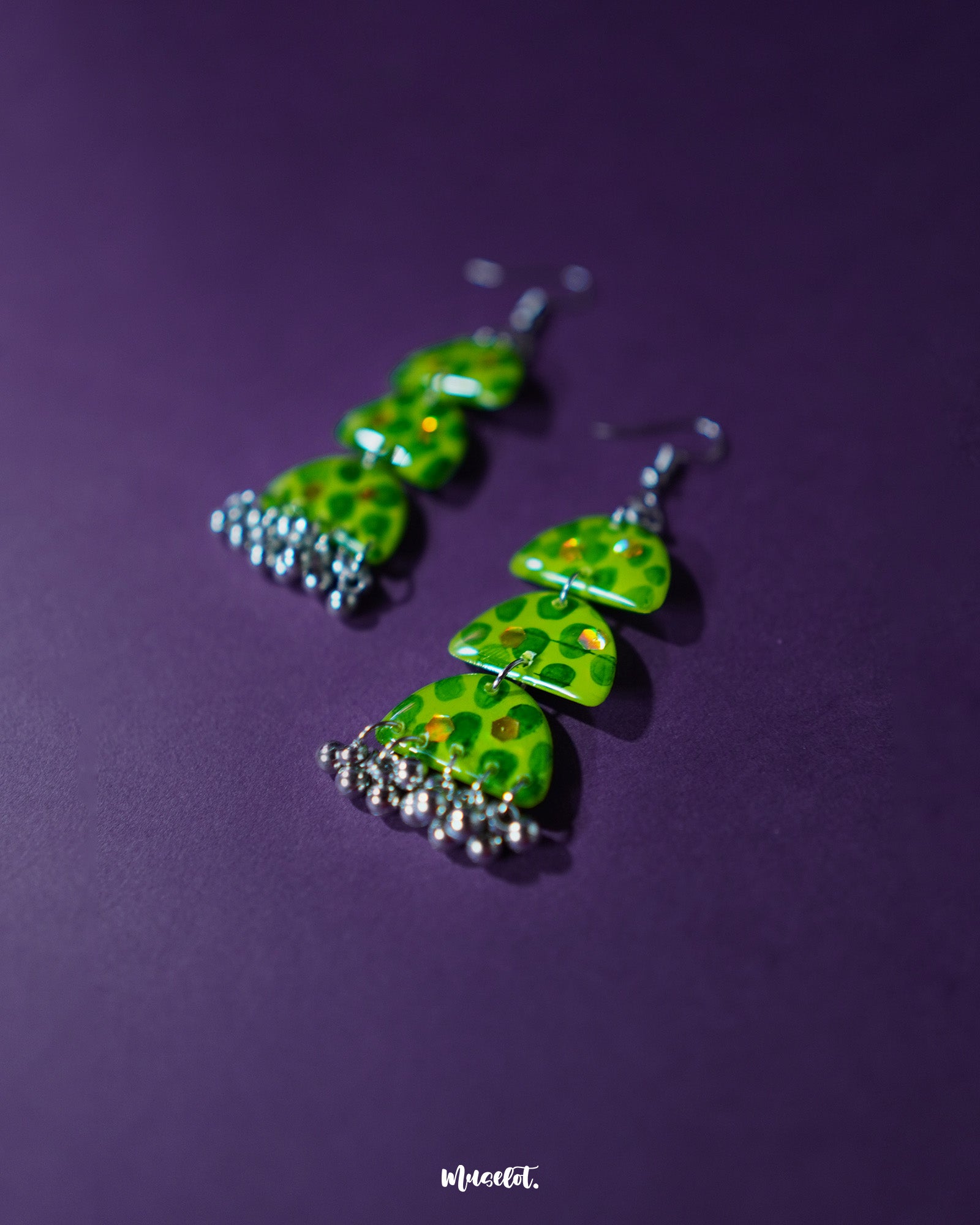 A pair of Parvaaz green drop earrings placed against a soft purple background, showcasing the elegant shape and subtle dark green dotted on a light green base detailing that add a touch of earthy grace — Muselot.