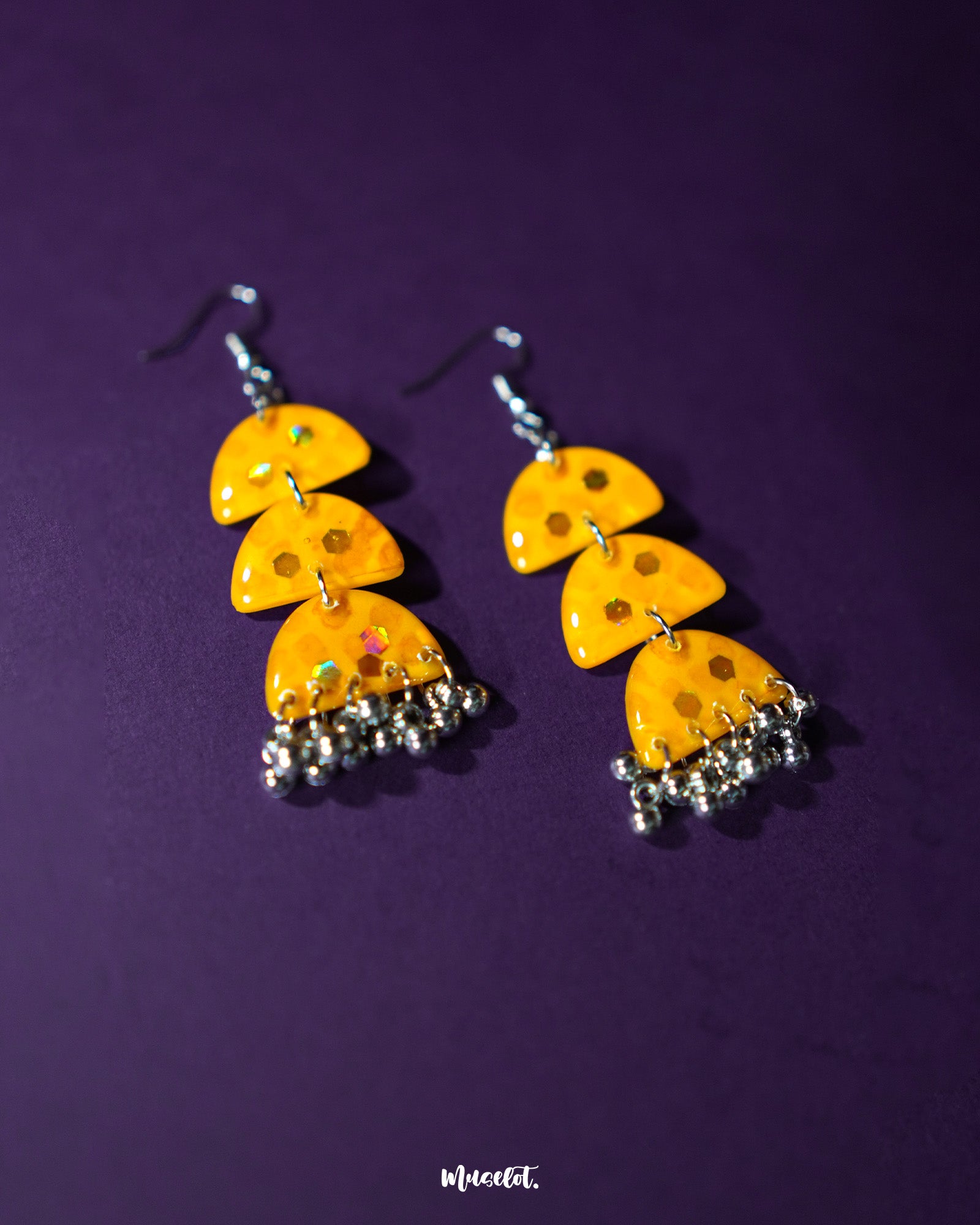 A pair of Parvaaz yellow drop earrings placed against a soft purple background, highlighting their vibrant floral design and showcasing the delicate handcrafted details that bring a playful charm — Muselot.