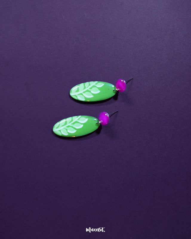 Patbaan drop earrings photographed against a rich purple background, highlighting their green hand-painted leaf design and vibrant pink studs — a striking contrast that enhances their artisanal elegance — Muselot.