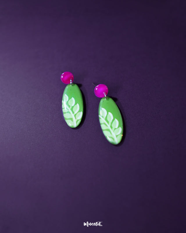 Close-up of the Patbaan drop earrings featuring delicate hand-painted white leaf motifs on an oval green base, paired with bold pink studs for a lively, nature-inspired statement — Muselot.