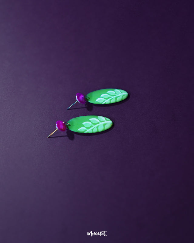 Patbaan drop earrings displayed in full view, showcasing their glossy green base adorned with soft white leaf strokes and contrasting fuchsia pink studs, blending earthy charm with playful color pops — Muselot.