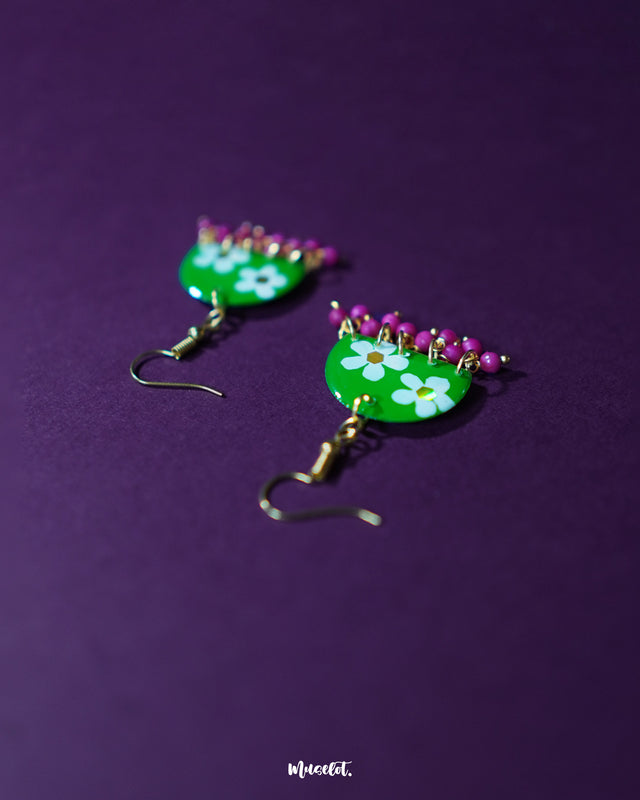 Close-up of Phoolbaan floral hook earrings featuring a rich green base with delicate white floral patterns and magenta bead accents, highlighting the fine handcrafted work and showcasing bold festive charm — Muselot.