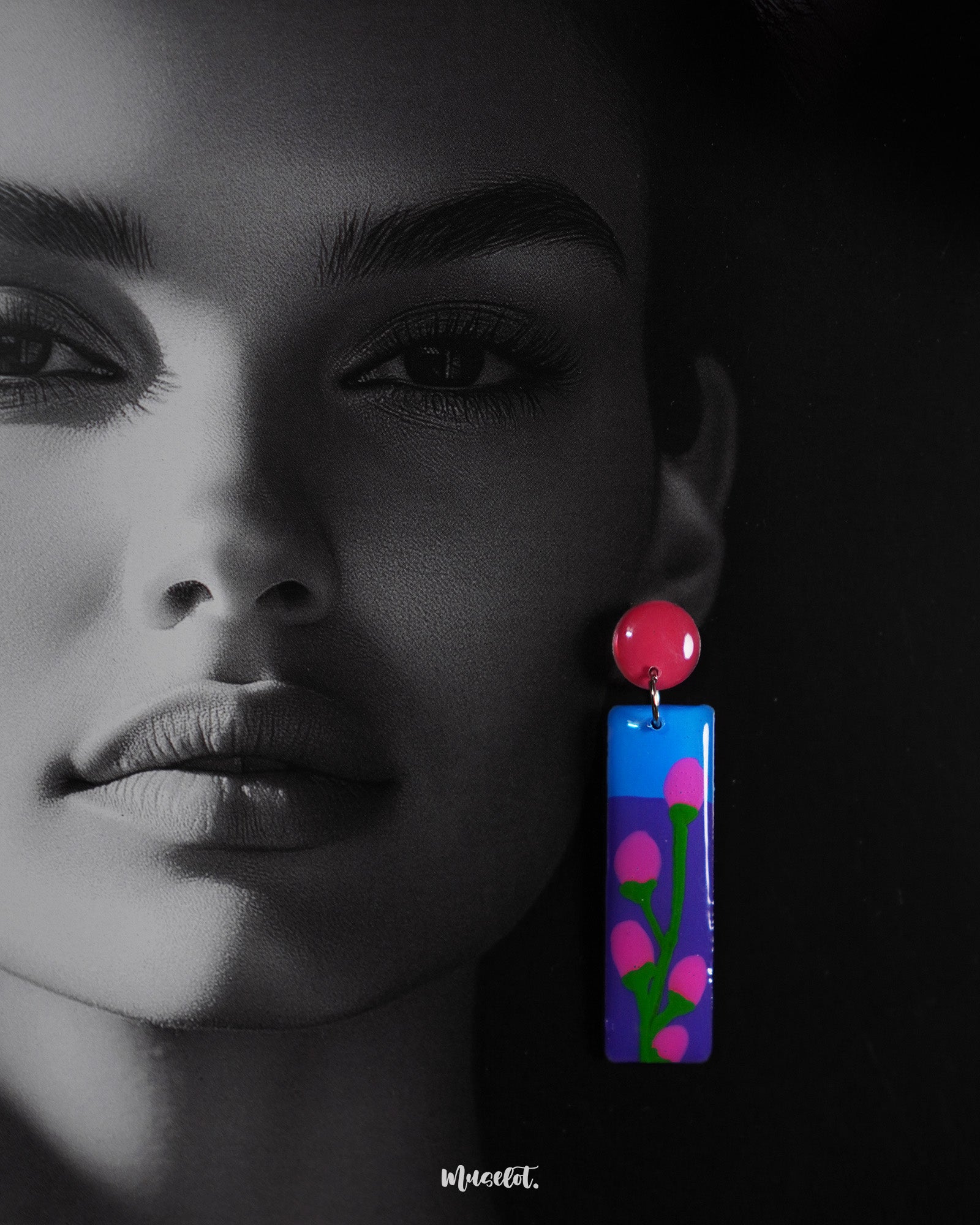A close-up of a woman wearing the Raatgul drop earrings highlights their glossy, hand-painted design. The deep blue and violet panels feature delicate pink buds with green stems, anchored by a bold red stud, capturing the essence of handcrafted elegance — Muselot.