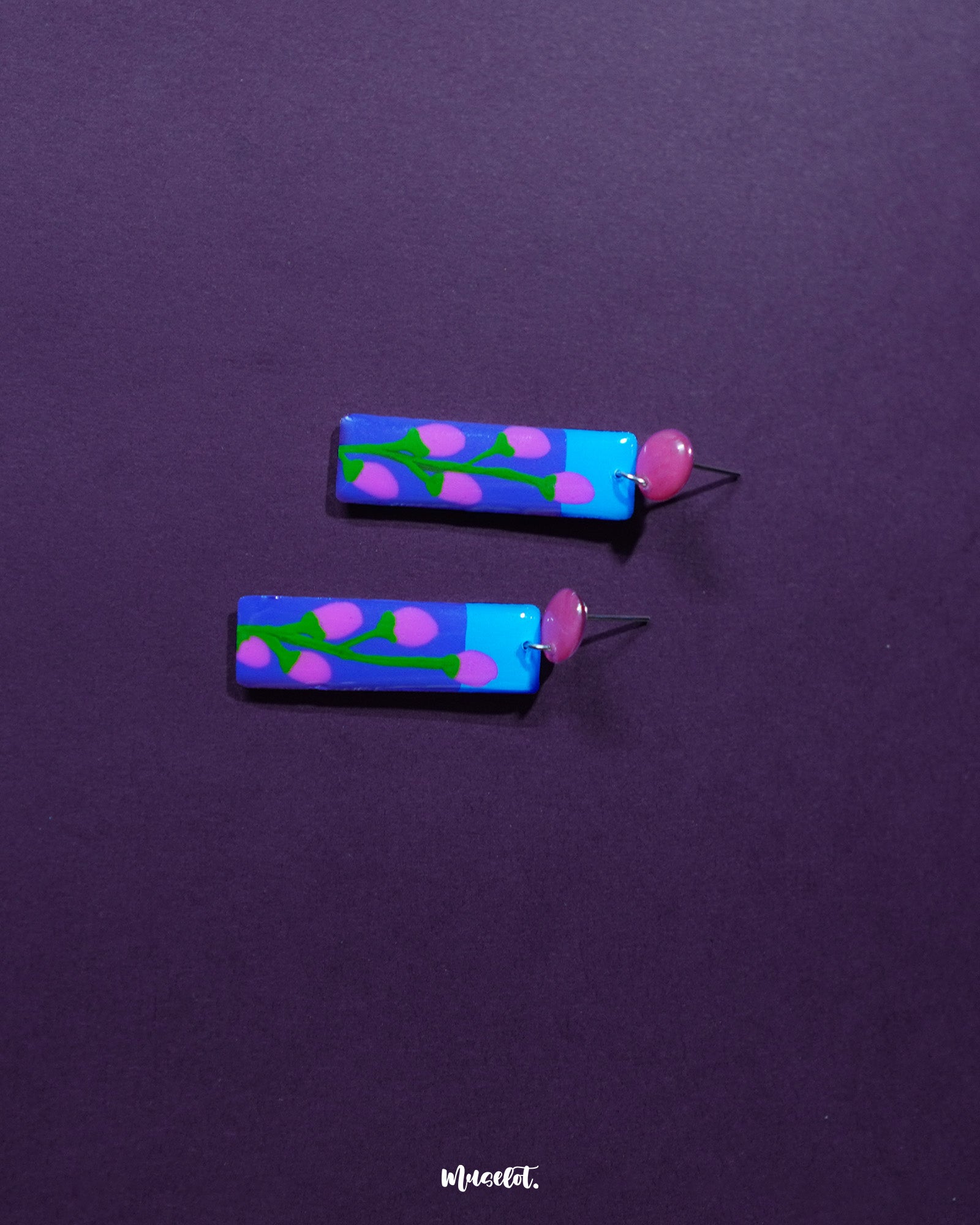 The Raatgul floral earrings rest against a rich purple backdrop, bringing out the vibrancy of their hand-painted details. With red studs and a striking blue-violet drop adorned with soft pink buds and green stems, these earrings evoke the charm of a midnight garden in bloom — Muselot.