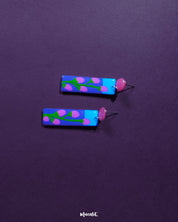 The Raatgul floral earrings rest against a rich purple backdrop, bringing out the vibrancy of their hand-painted details. With red studs and a striking blue-violet drop adorned with soft pink buds and green stems, these earrings evoke the charm of a midnight garden in bloom — Muselot.
