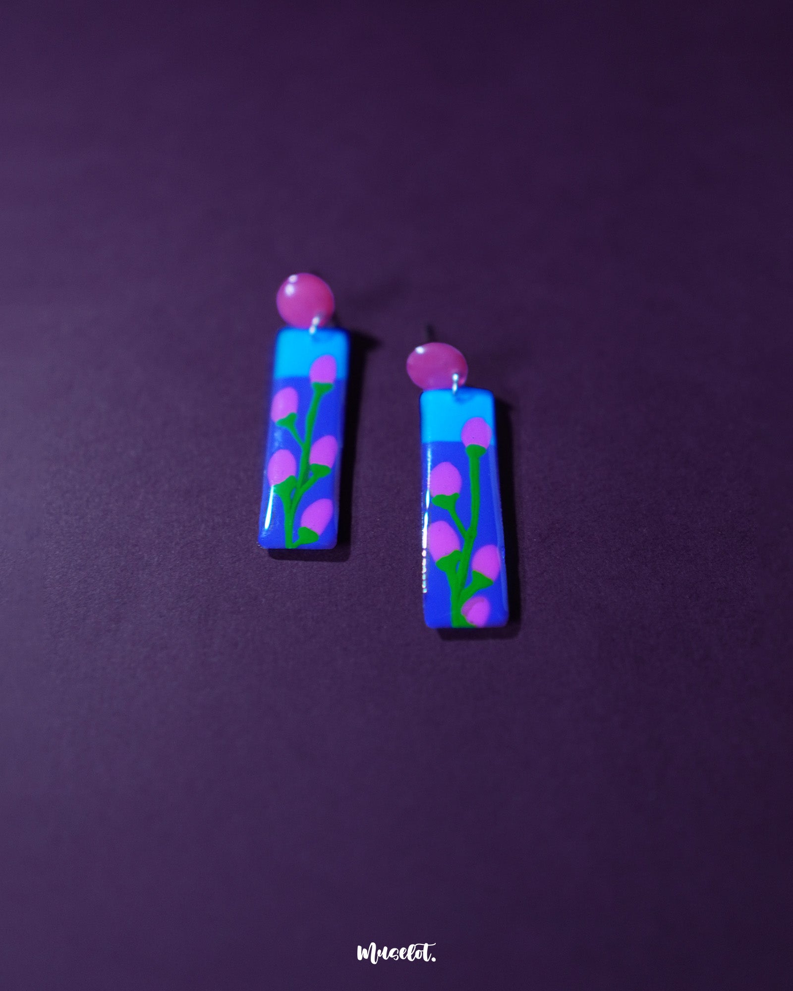 A striking pair of hand-painted earrings featuring a vivid blue and violet base adorned with delicate pink floral buds and green stems. The glossy finish and red circular studs add a playful yet artistic flair — Muselot.