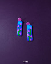 A striking pair of hand-painted earrings featuring a vivid blue and violet base adorned with delicate pink floral buds and green stems. The glossy finish and red circular studs add a playful yet artistic flair — Muselot.