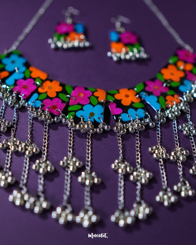 A beautifully arranged flat lay of the Rangbaagh black floral necklace set featuring vibrant floral accents and metallic gungroo elements, laid out against a soft purple backdrop to showcase its handcrafted detail and festive allure — Muselot