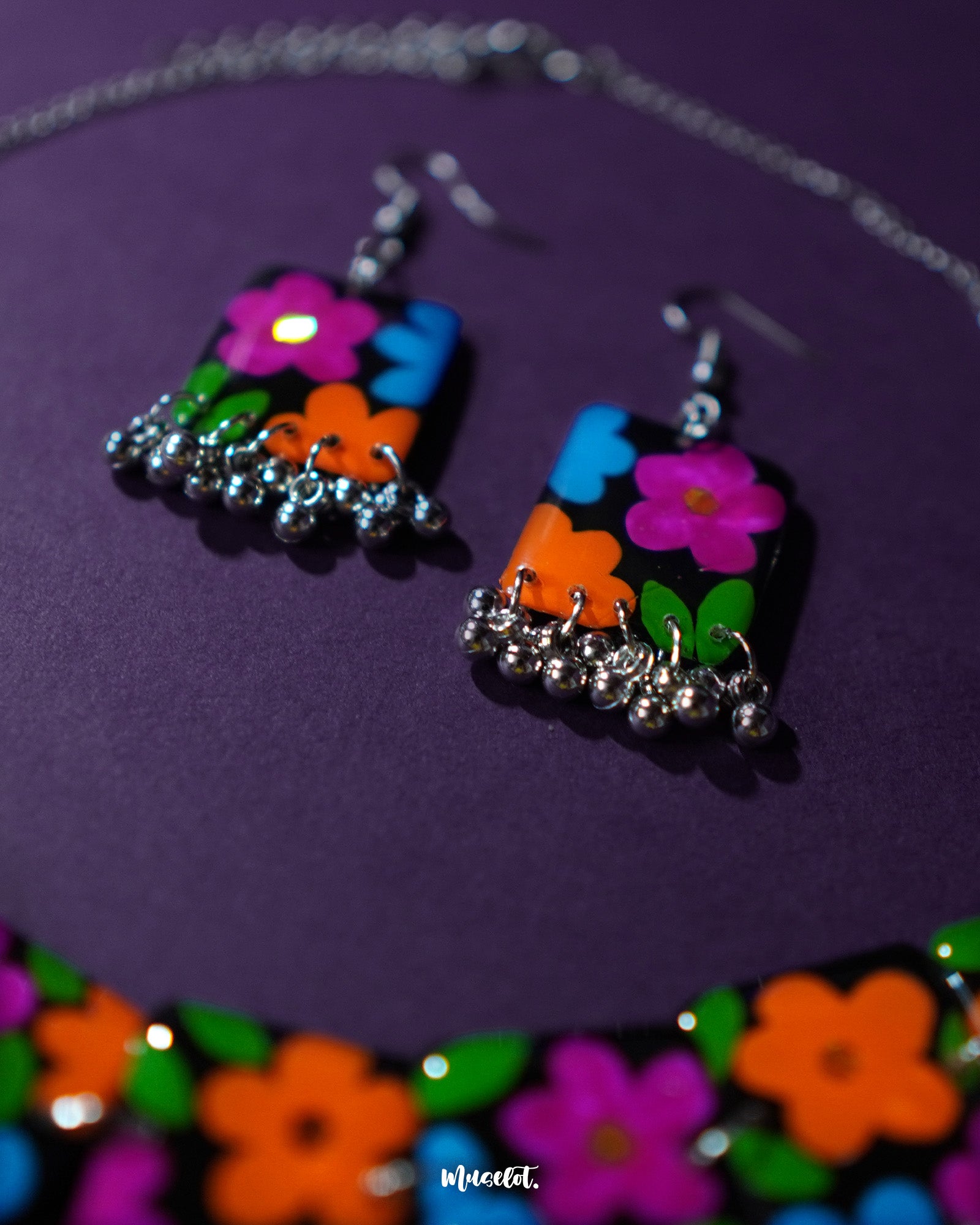 A close-up view of the Rangbaagh black floral earrings featuring delicate blue, orange, and purple flowers arranged in a balanced composition, paired with metallic gungroo drops that add a festive jingle and artisanal charm — Muselot