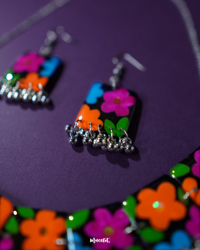 A close-up of the earrings from the Rangbaagh black floral necklace set, featuring pink and orange floral motifs suspended from a delicate gunghroo-accented base, offering a bold yet graceful statement — Muselot.