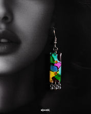 Close-up of a model wearing a vibrant rectangular earring from the Rangbaari colorful jewelry set by Muselot. The earring features a blend of bright green, pink, yellow, and blue hues, accented with small silver ghungroos at the bottom, standing out against the model’s dark hair and skin.