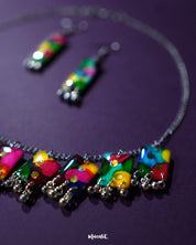 Rangbaari colorful jewelry set with a silver chain necklace and matching earrings, featuring bold multicolor abstract rectangular charms and delicate silver bead accents, artistically displayed on a deep purple background – Muselot.