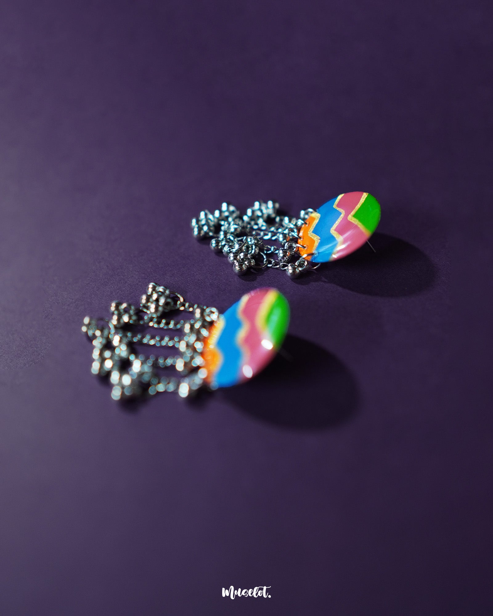 A pair of Rangtarang colorful long chain earrings displayed against a rich purple background, featuring bold zigzag patterns in green, pink, blue, and orange with dangling silver chains and clustered beads that bring a festive, boho vibe – Muselot.