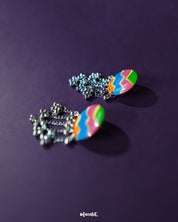 A pair of Rangtarang colorful long chain earrings displayed against a rich purple background, featuring bold zigzag patterns in green, pink, blue, and orange with dangling silver chains and clustered beads that bring a festive, boho vibe – Muselot.
