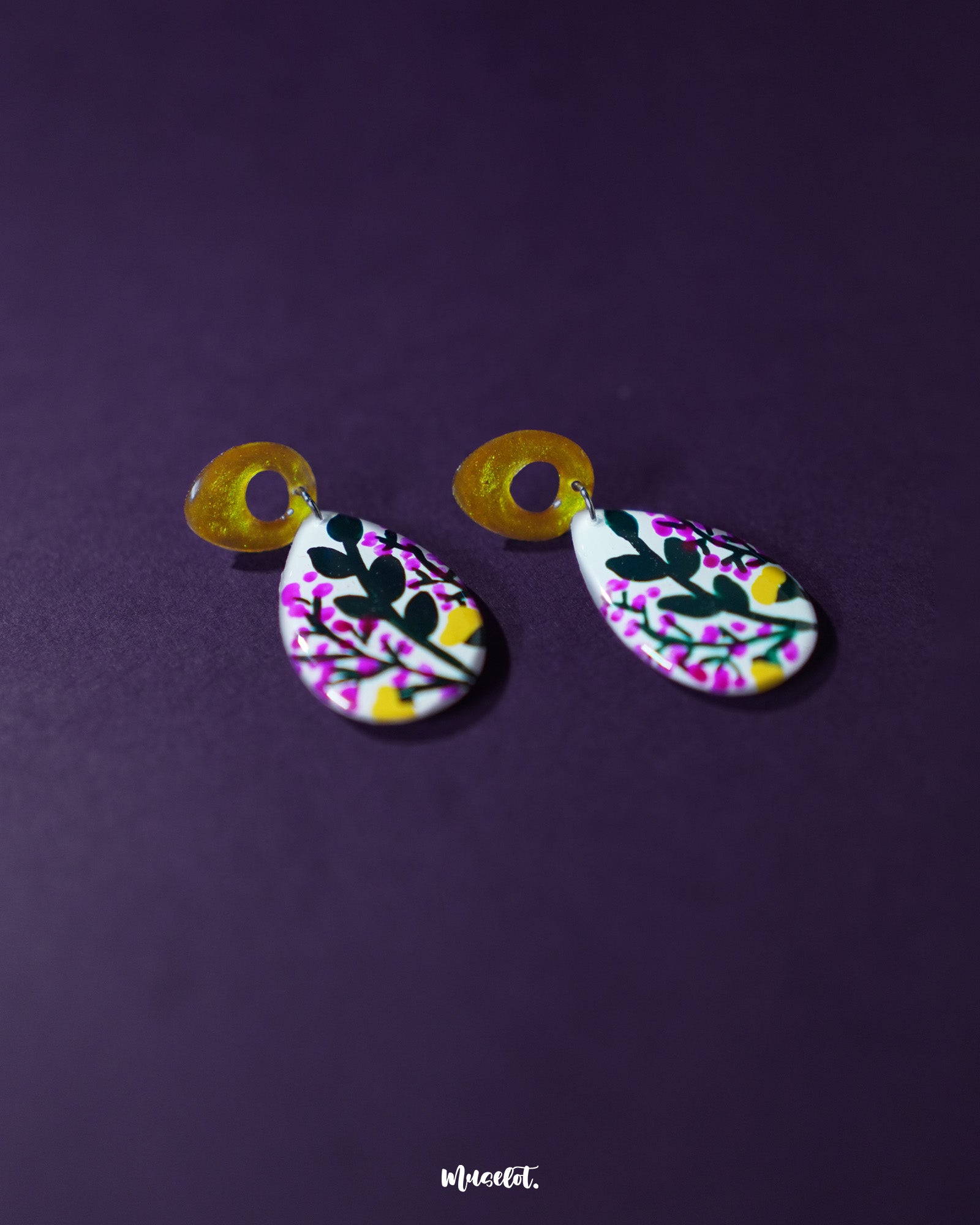 Raushanbahaar floral drop earrings resting on a deep purple background, showcasing teardrop-shaped dangles painted with pink buds, green leaves, and yellow accents, attached to shimmering golden open-loop studs — Muselot.