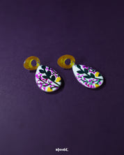 Raushanbahaar floral drop earrings resting on a deep purple background, showcasing teardrop-shaped dangles painted with pink buds, green leaves, and yellow accents, attached to shimmering golden open-loop studs — Muselot.