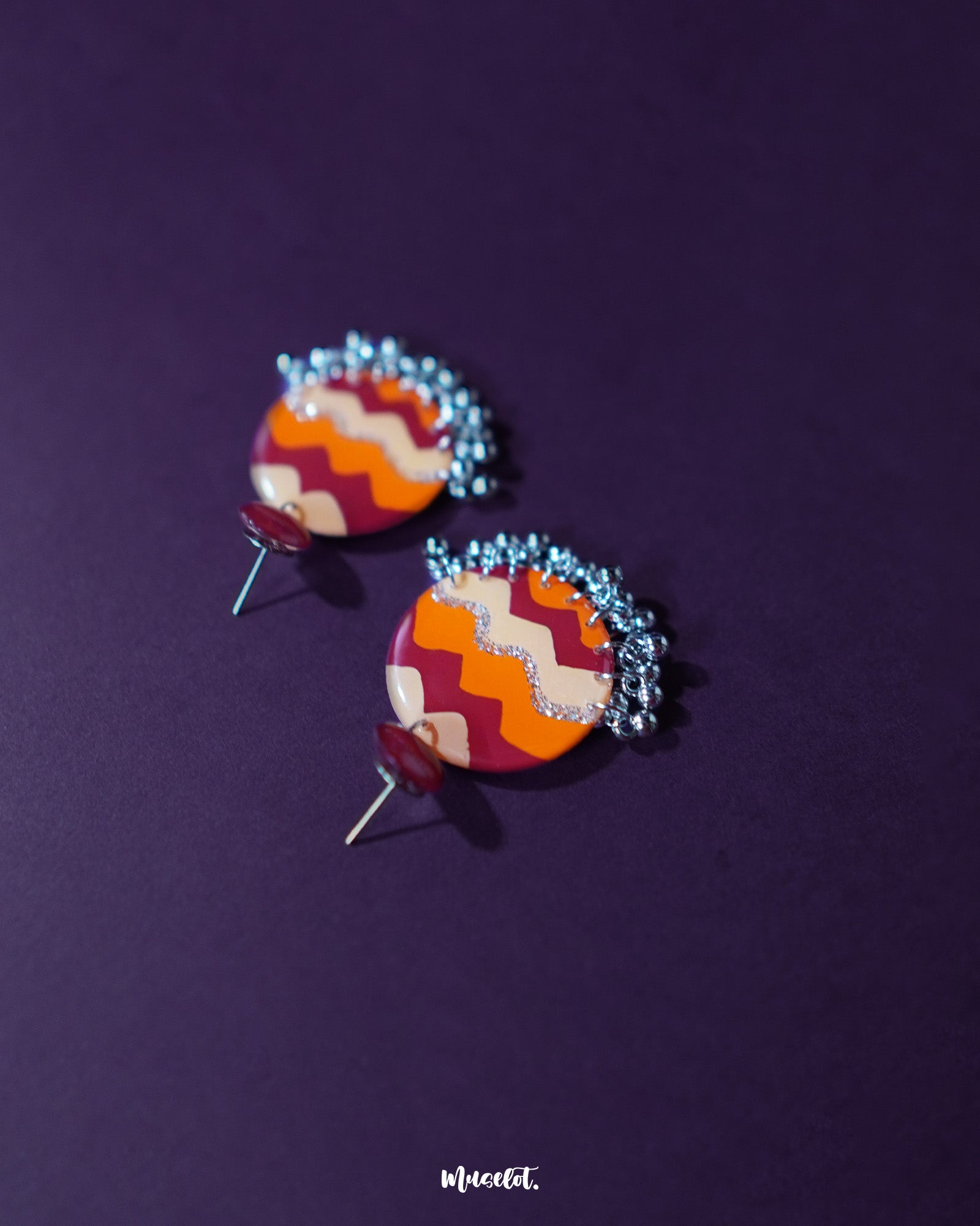 Sahar traditional stud earrings rest elegantly against a deep purple backdrop, showcasing their bold round form painted with maroon, orange, and mustard waves, finished with delicate silver ghungroos that lend a festive, handcrafted touch — Muselot.