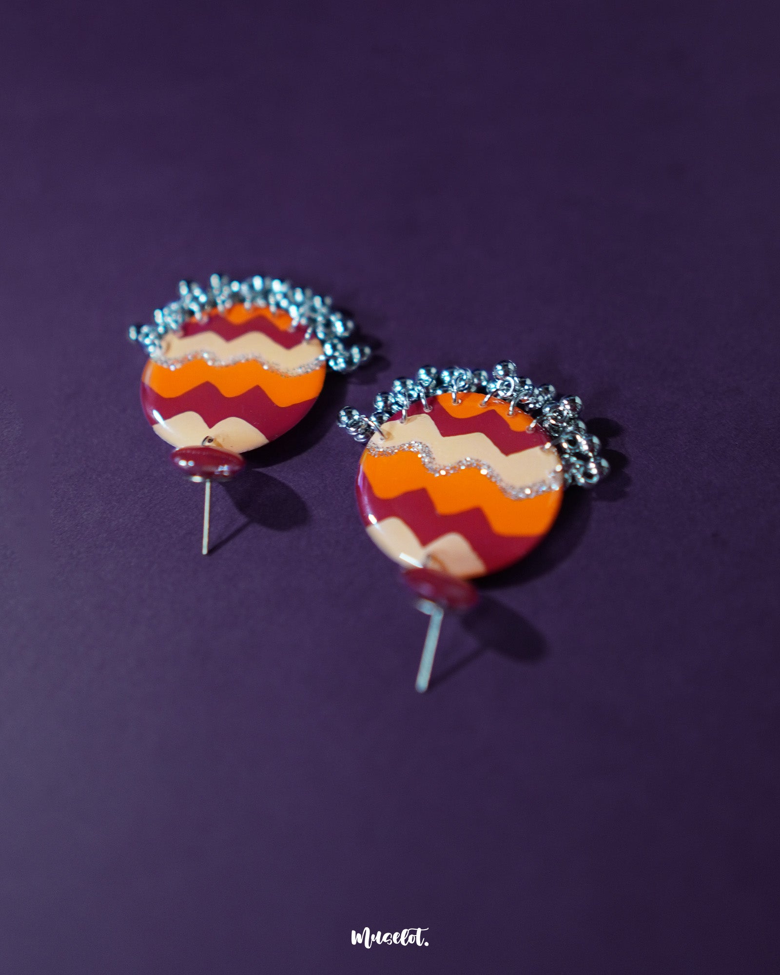A pair of Sahar traditional stud earrings displayed flat, featuring bold concentric waves in maroon, orange, mustard, and glittering silver, with tiny silver ghungroos adding a rhythmic charm to the vibrant hand-painted design — Muselot.