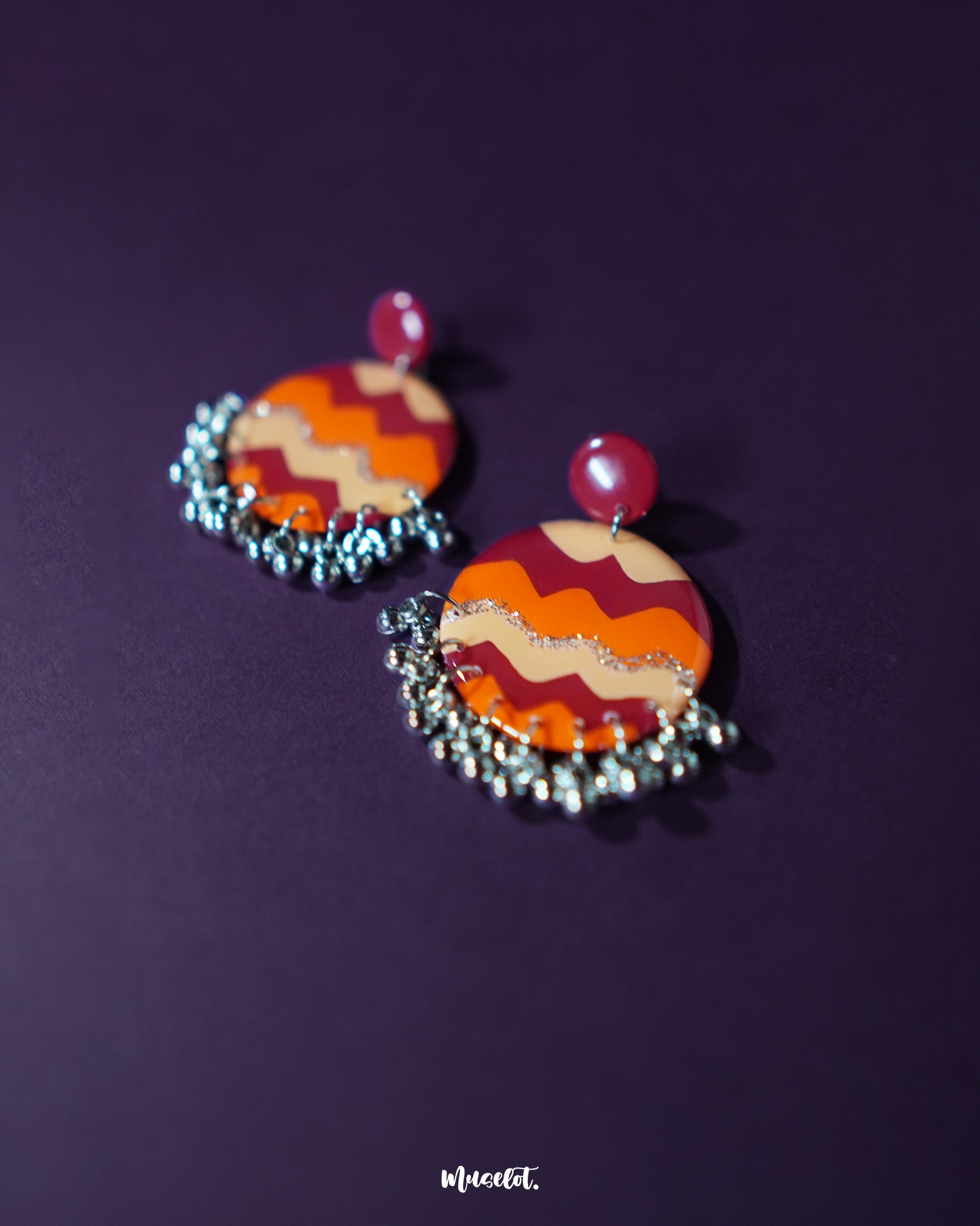 Close-up of the Sahar traditional stud earrings resting on textured fabric, showcasing the intricate hand-painted circular pattern in deep maroon, orange, mustard, and shimmering silver, accented with tiny silver ghungroos that lend a festive and artisanal appeal — Muselot.