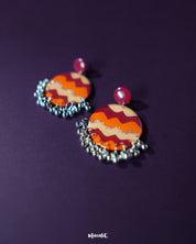 Close-up of the Sahar traditional stud earrings resting on textured fabric, showcasing the intricate hand-painted circular pattern in deep maroon, orange, mustard, and shimmering silver, accented with tiny silver ghungroos that lend a festive and artisanal appeal — Muselot.