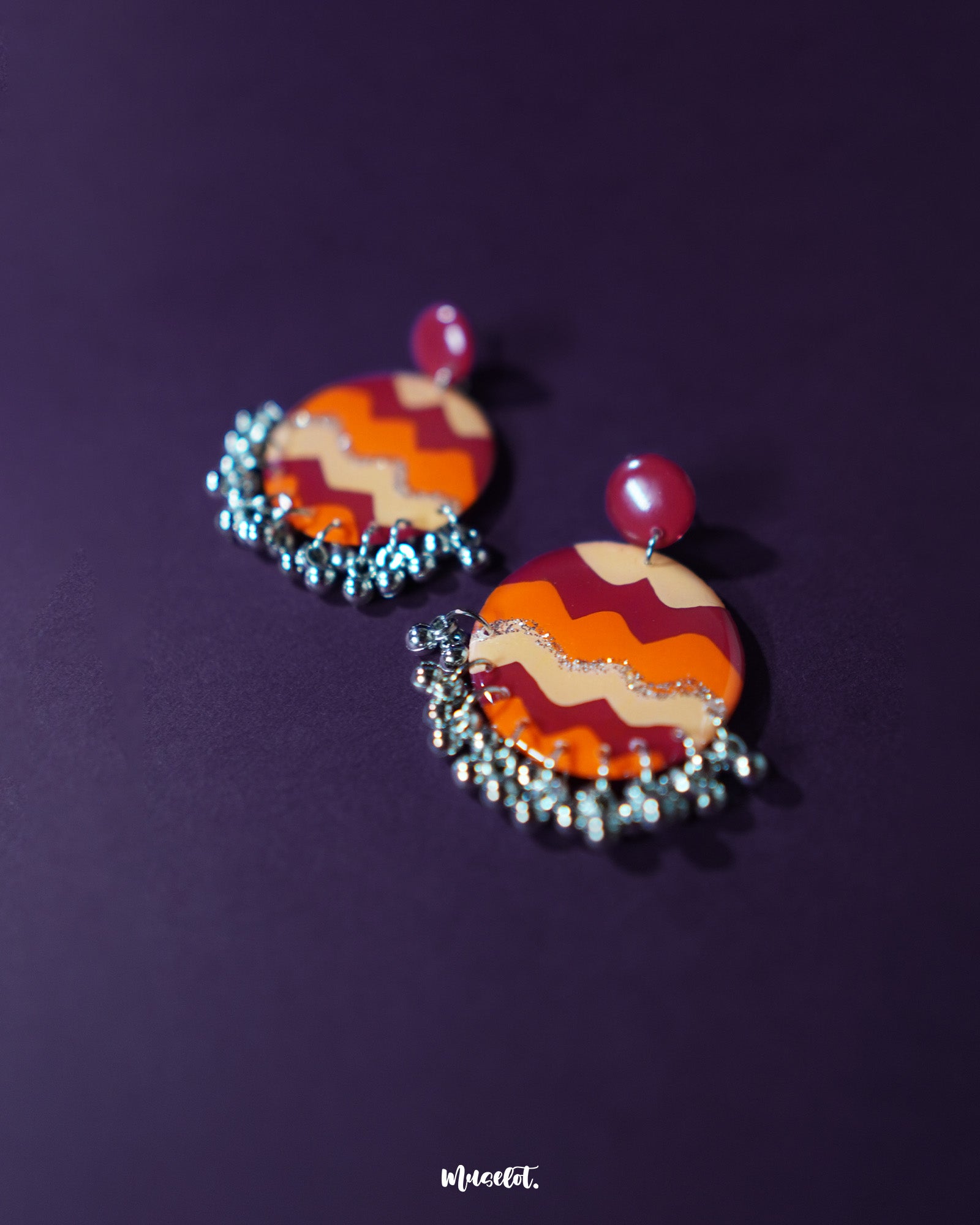 Flat lay of the Sahar traditional stud earrings placed on an orange backdrop, highlighting their bold circular design with earthy tones of mustard, rust, and silver, finished with delicate silver ghungroos that dangle subtly — Muselot.