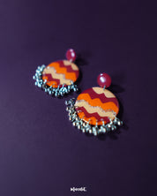 Flat lay of the Sahar traditional stud earrings placed on an orange backdrop, highlighting their bold circular design with earthy tones of mustard, rust, and silver, finished with delicate silver ghungroos that dangle subtly — Muselot.