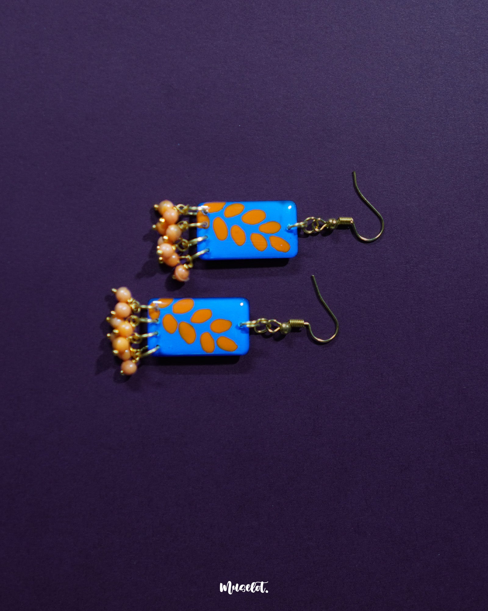 A pair of Sandhyaraag hook floral earrings resting on a purple background, showcasing a vivid blue base with scattered orange petal designs and golden hooks, finished with orange beaded danglers for a playful boho vibe — Muselot.
