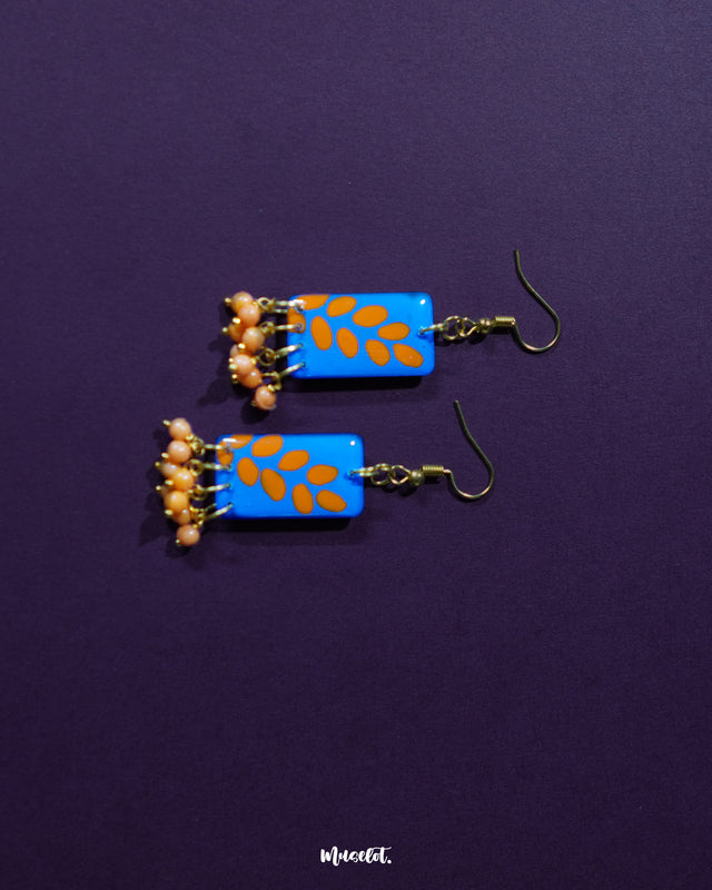 A pair of Sandhyaraag hook floral earrings resting on a purple background, showcasing a vivid blue base with scattered orange petal designs and golden hooks, finished with orange beaded danglers for a playful boho vibe — Muselot.