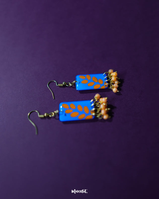 Close-up of Sandhyaraag hook floral earrings showcasing vibrant orange bead danglers against a bright blue base with scattered floral patterns, highlighting the lively boho vibe and detailed hand-painted design — Muselot.