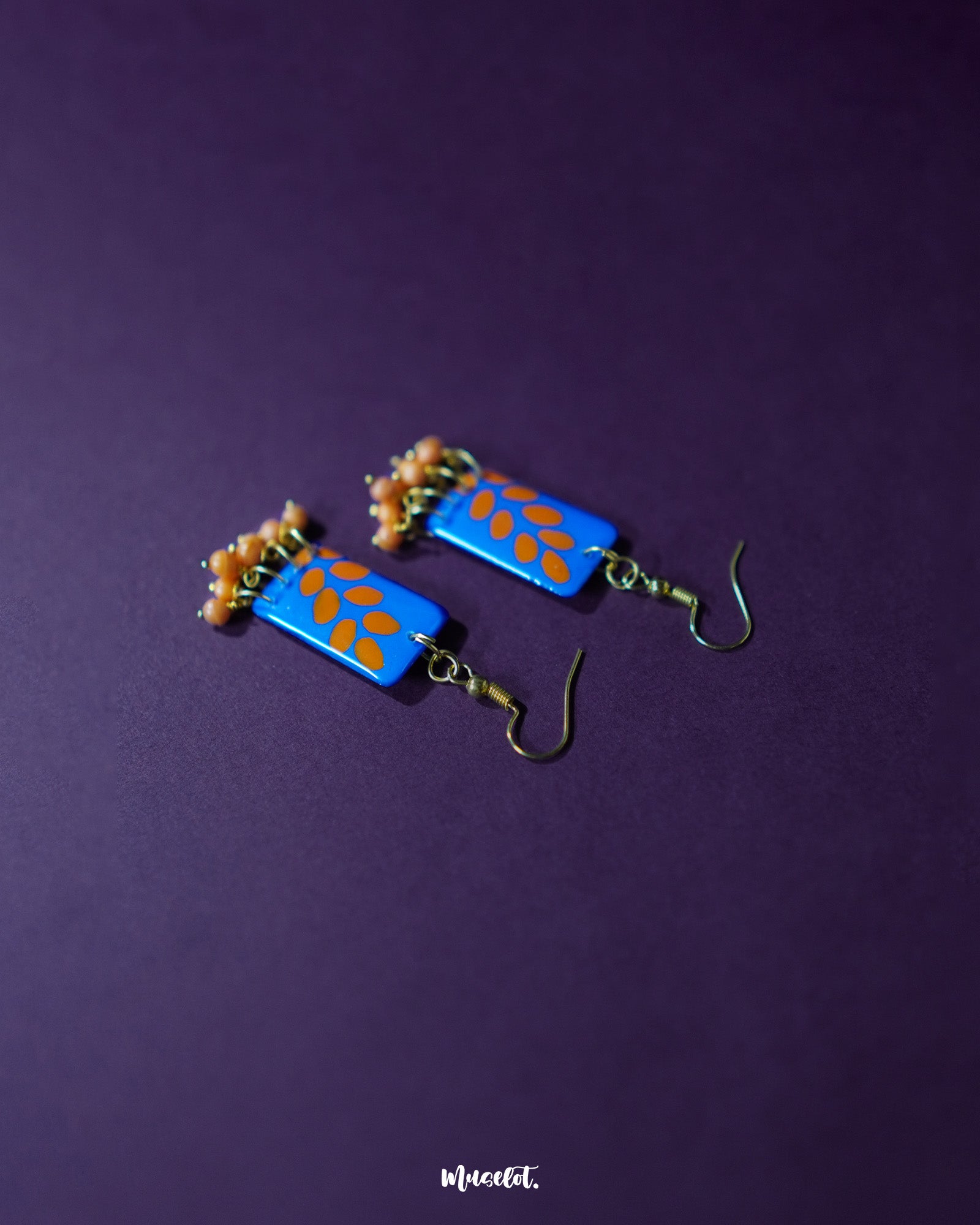 A pair of Sandhyaraag hook floral earrings placed against a rich purple backdrop, showcasing their vibrant blue and orange palette with delicate floral detailing and handcrafted charm — Muselot.