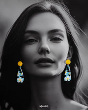 A model wearing the Shaamgul handmade drop earrings showcases their sleek, elongated shape adorned with hand-painted white flowers and black leaves on a pastel blue base, highlighting the bold pop of yellow from the round studs for a vibrant, artistic finish — Muselot.