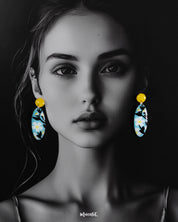 A woman wears the oval-shaped Shaamgul floral drop earrings against a deep purple background, highlighting the sky-blue base, white daisies, black leaves, and yellow stud—beautifully showcasing their handmade charm and standout design — Muselot.