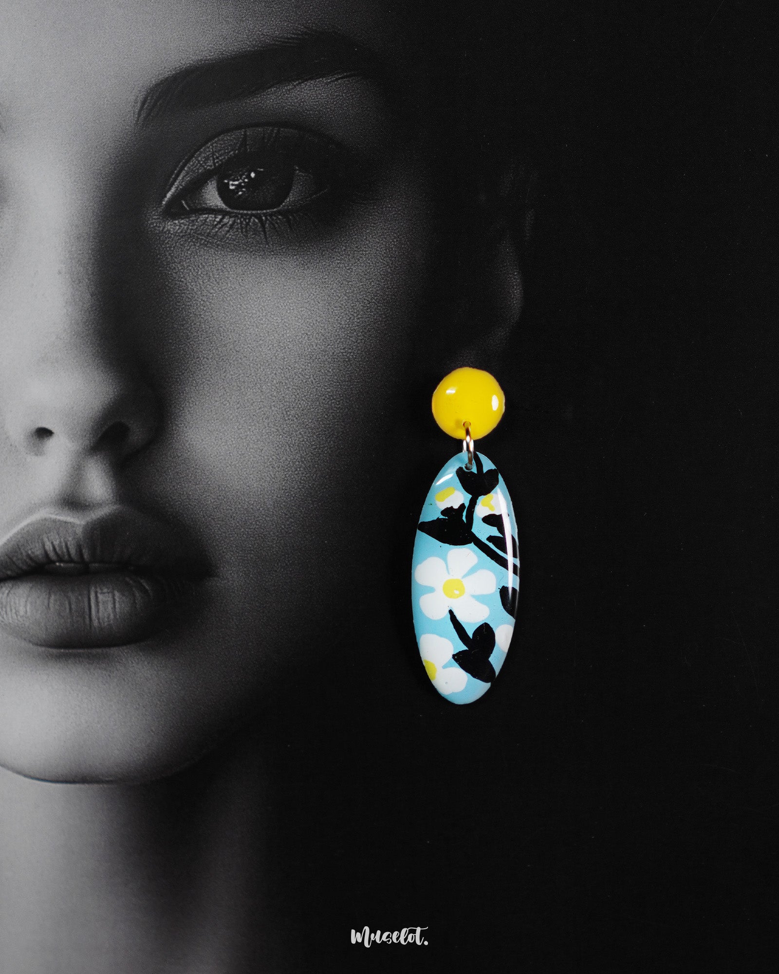 Close-up of a woman wearing the oval-shaped Shaamgul floral drop earrings, set against a rich purple background, showcasing the sky-blue base, hand-painted white daisies, black leafy details, and yellow stud for a striking, handmade statement — Muselot.