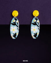Oval-shaped Shaamgul floral drop earrings displayed against a rich purple background, featuring a sky-blue base with white daisy motifs, bold black leaves, and a striking yellow circular stud, creating a charming contrast — Muselot.