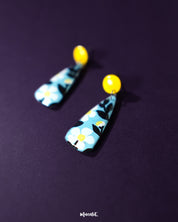 Shaamgul floral drop earrings resting on fabric, showcasing their unique shape with black and white floral accents and a soft blue drop, highlighting the handcrafted design — Muselot.