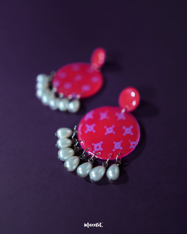 A closer look at the Shabnam pink pearl drop earrings placed on a soft beige background, showcasing the glossy finish of the round pink tops and the elegant white pearl-like drops hanging below, highlighting their handcrafted finesse and graceful appeal — Muselot.